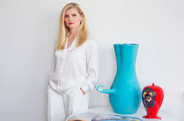 A blonde woman wearing a white shirt and white trousers standing next to a table with a blue vase and a red ornament