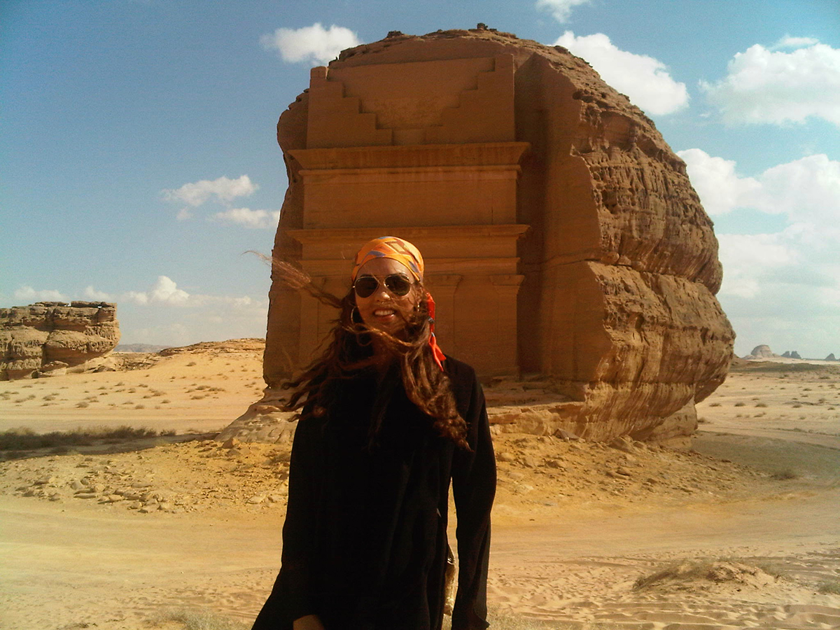  A woman wearing a black dress and orange head scarf standing next to a large rock in a desert