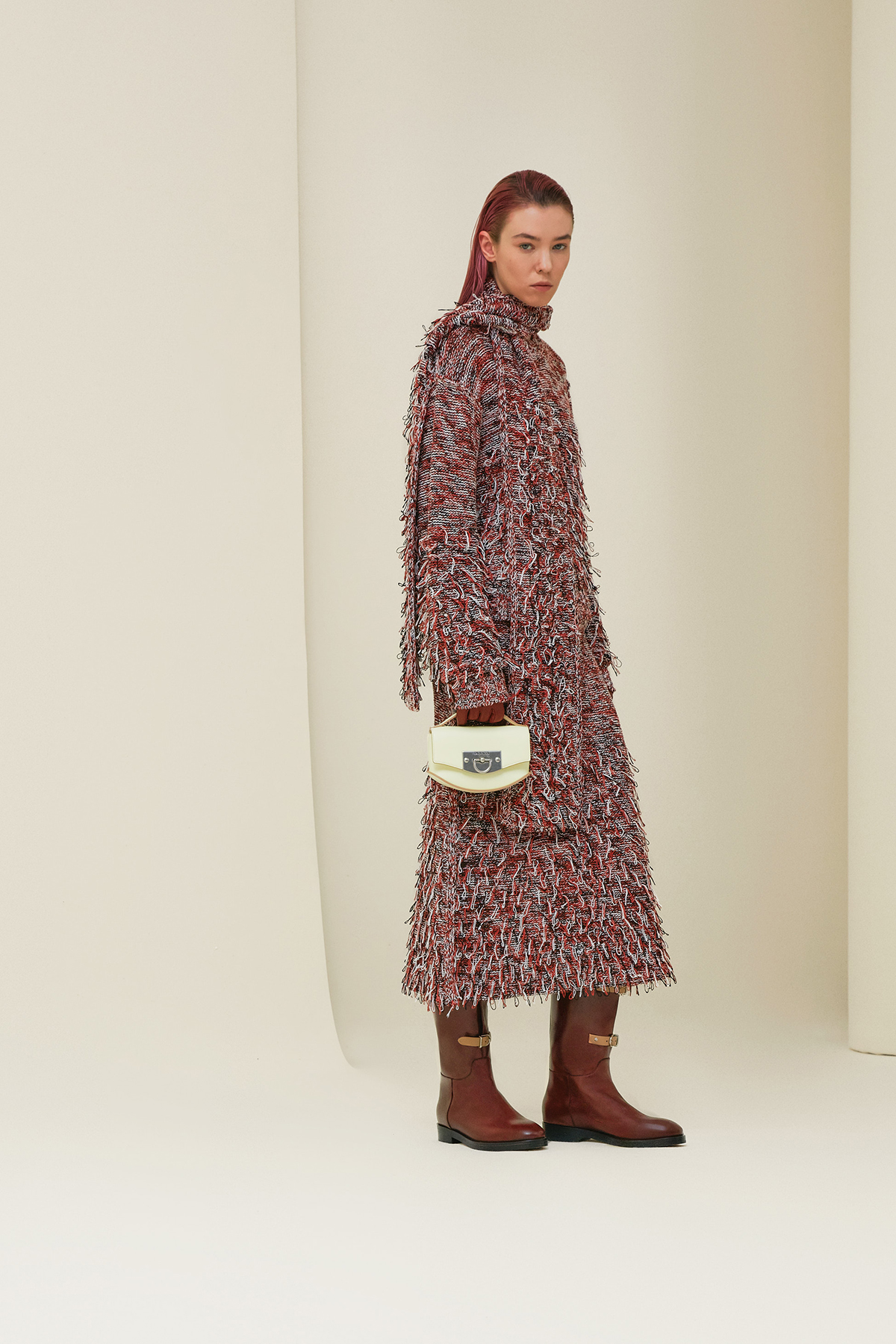 A woman wearing a tweed pink an red cot with red boots and holding a white bag