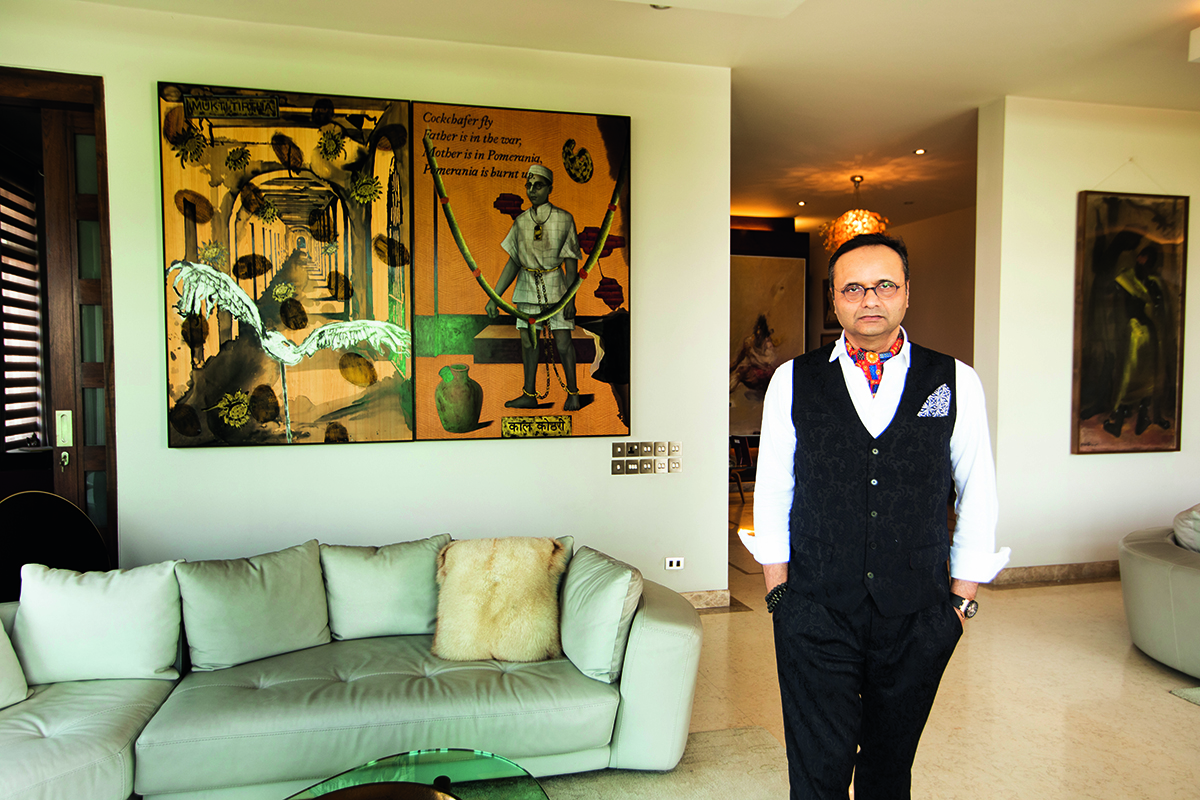 A man wearing a white shirt and black vest standing next to a green sofa and a large yellow painting behind him