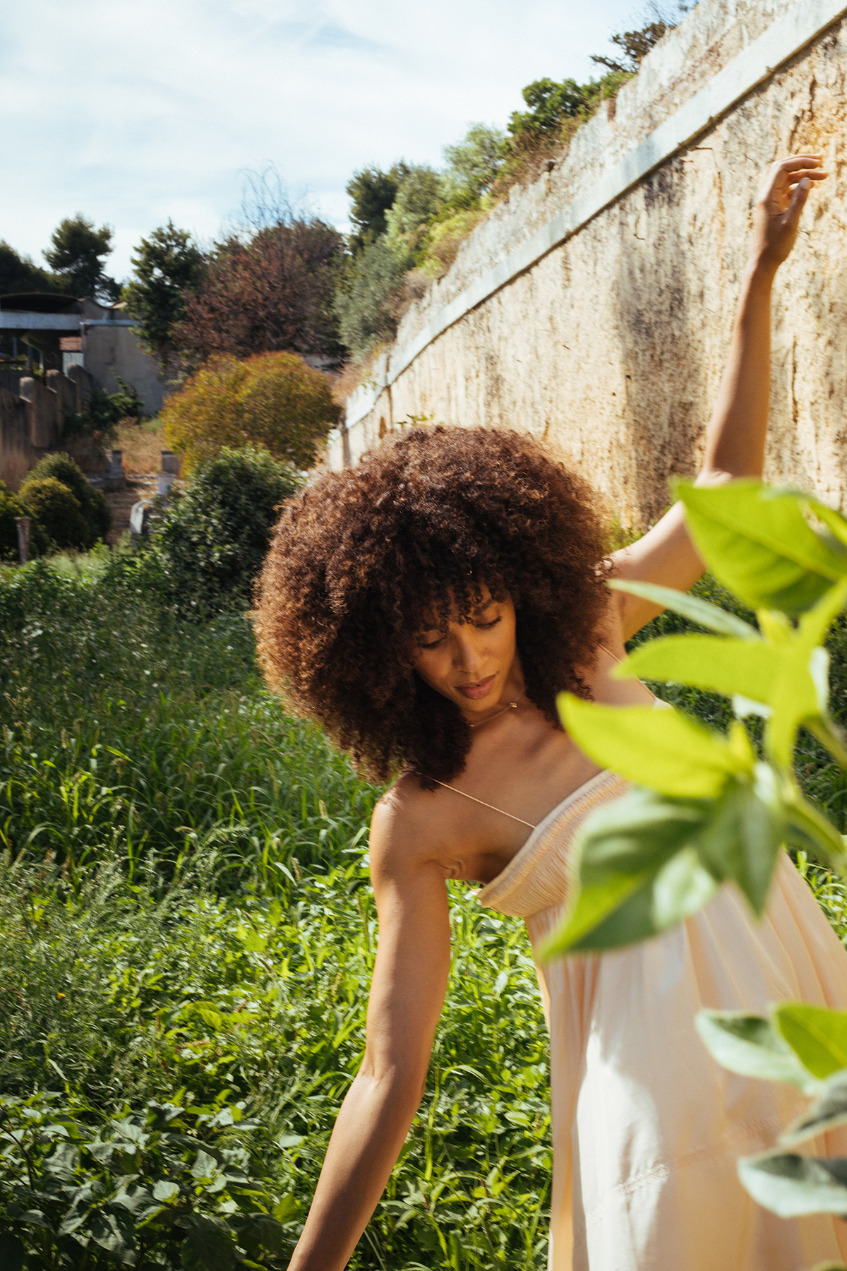 A woman standing in a garden holding onto a wall with one arm and plants around her