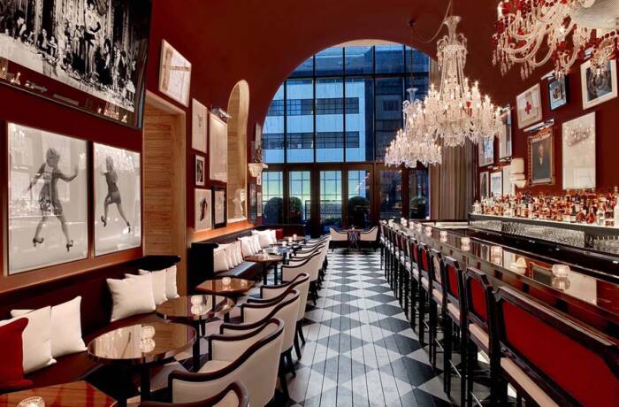 A red restaurant with a large window at the back and long rows of tables with benches and chairs on either side and crystal chandeliers over the bar